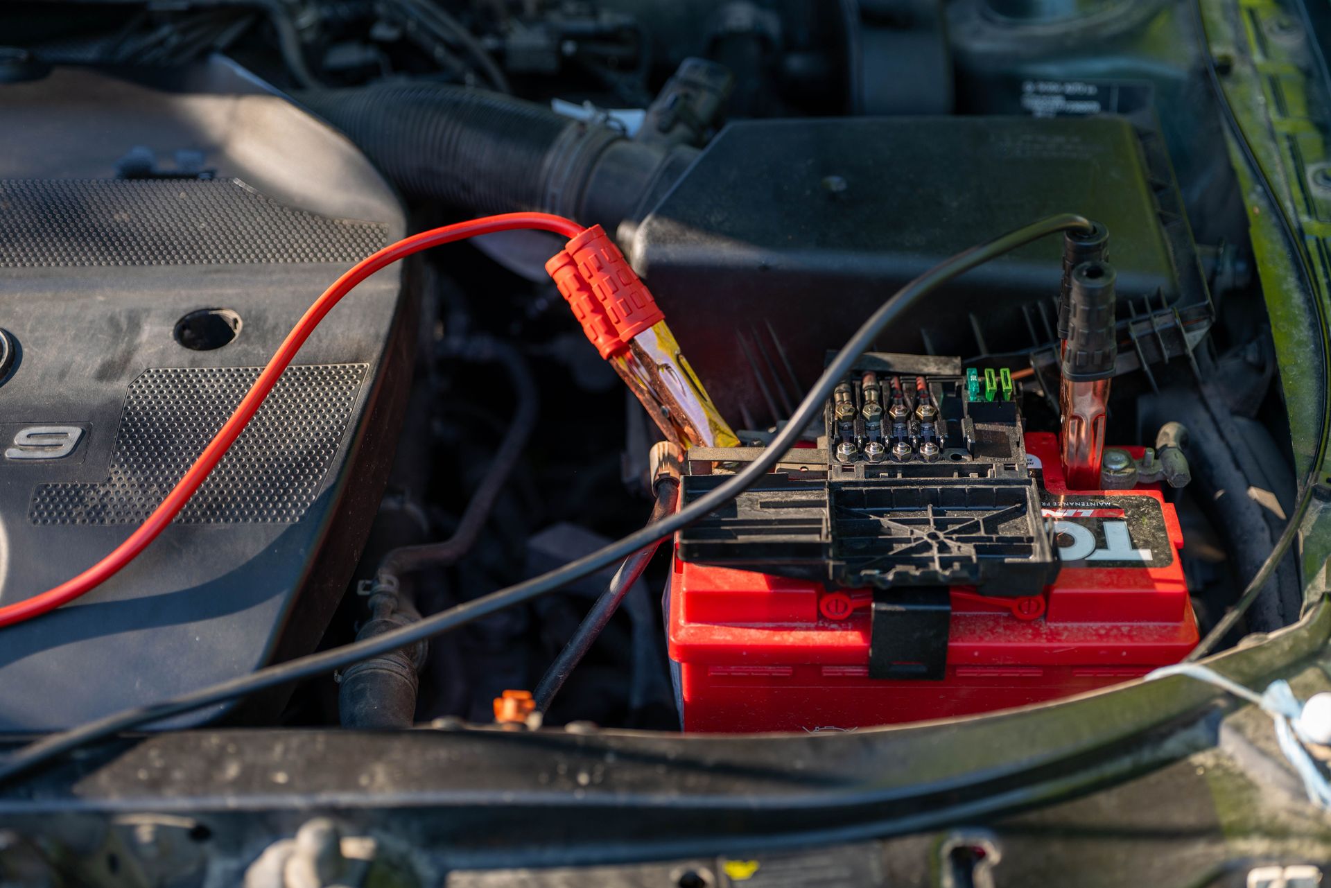 Close-up of a car battery with attached jumper cables in an engine bay.